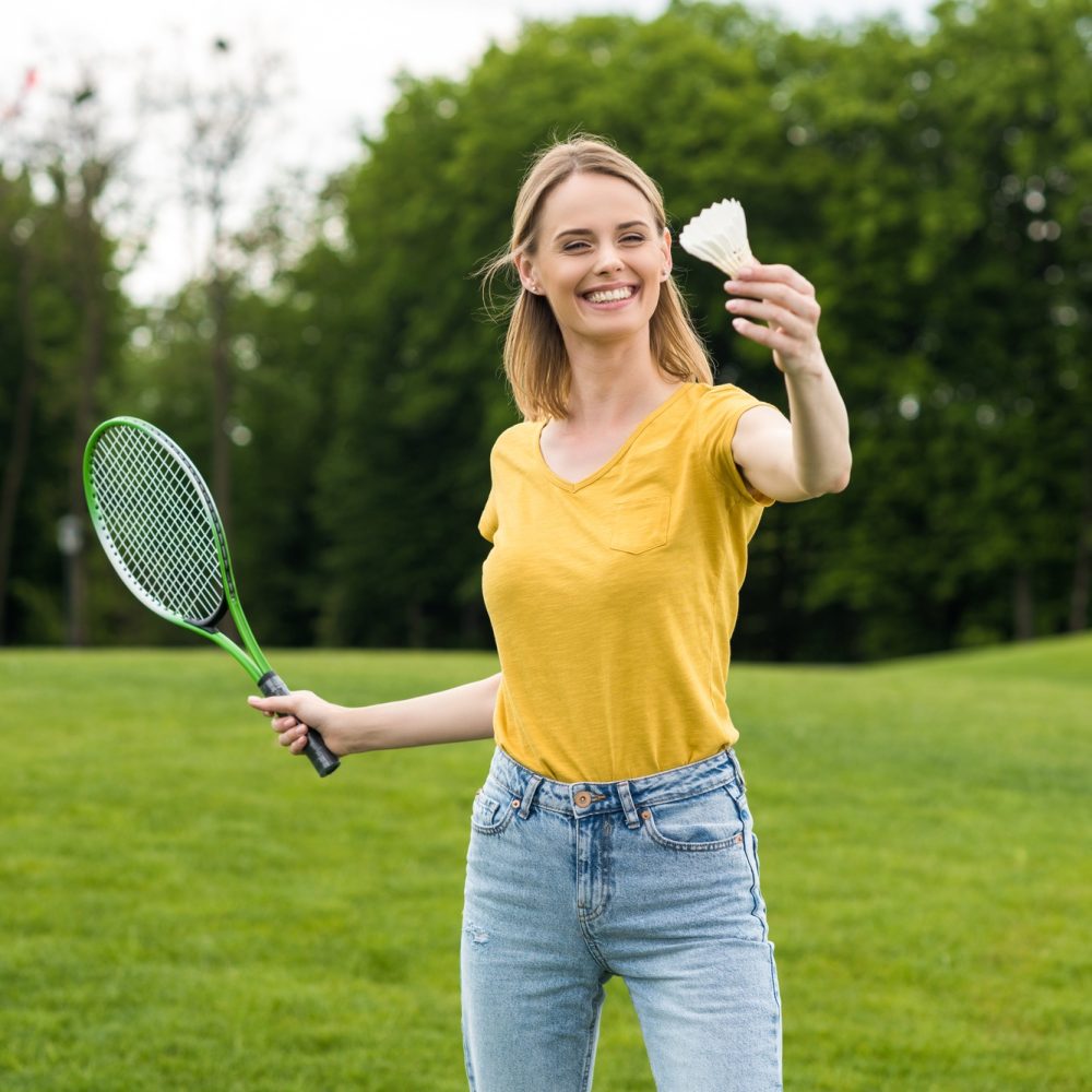 blonde smiling woman holding badminton racquet and shuttlecock in park